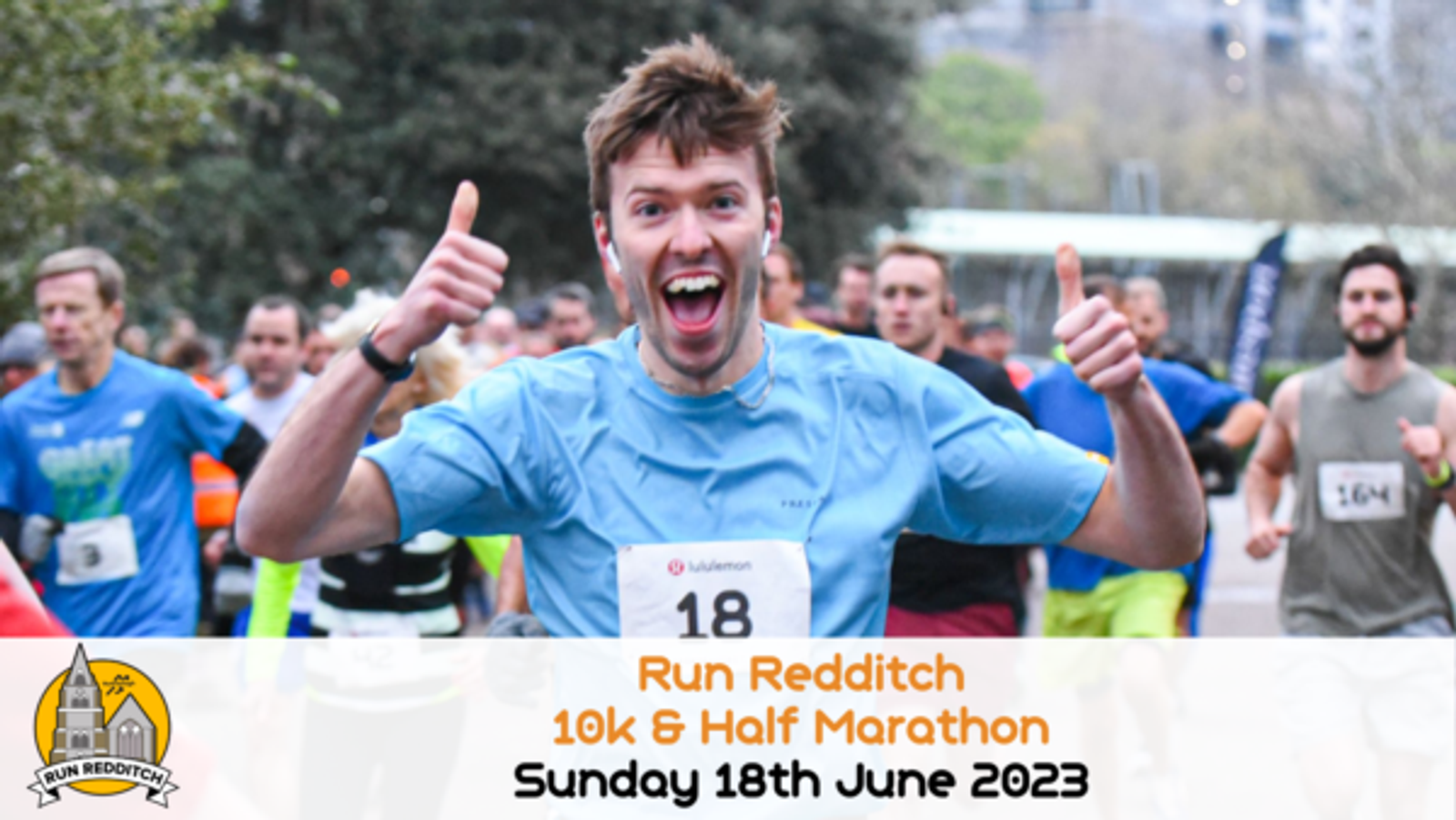 A runner in a blue shirt, numbered 18, is giving two thumbs up while running with a big smile. He is participating in the Run Redditch 10k & Half Marathon event on Sunday, 18th June 2023. Other runners can be seen in the background.
