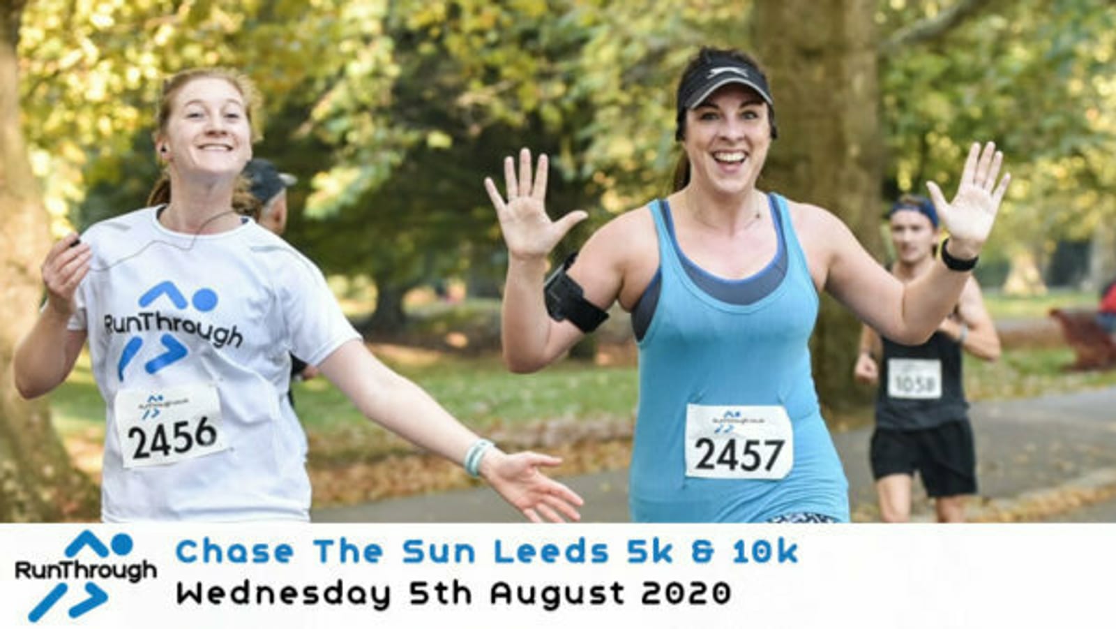 Two women smile and wave while running in a park during the Chase The Sun Leeds 5k & 10k event held on Wednesday, 5th August 2020. Other runners can be seen in the background. Both women are wearing RunThrough branded shirts.