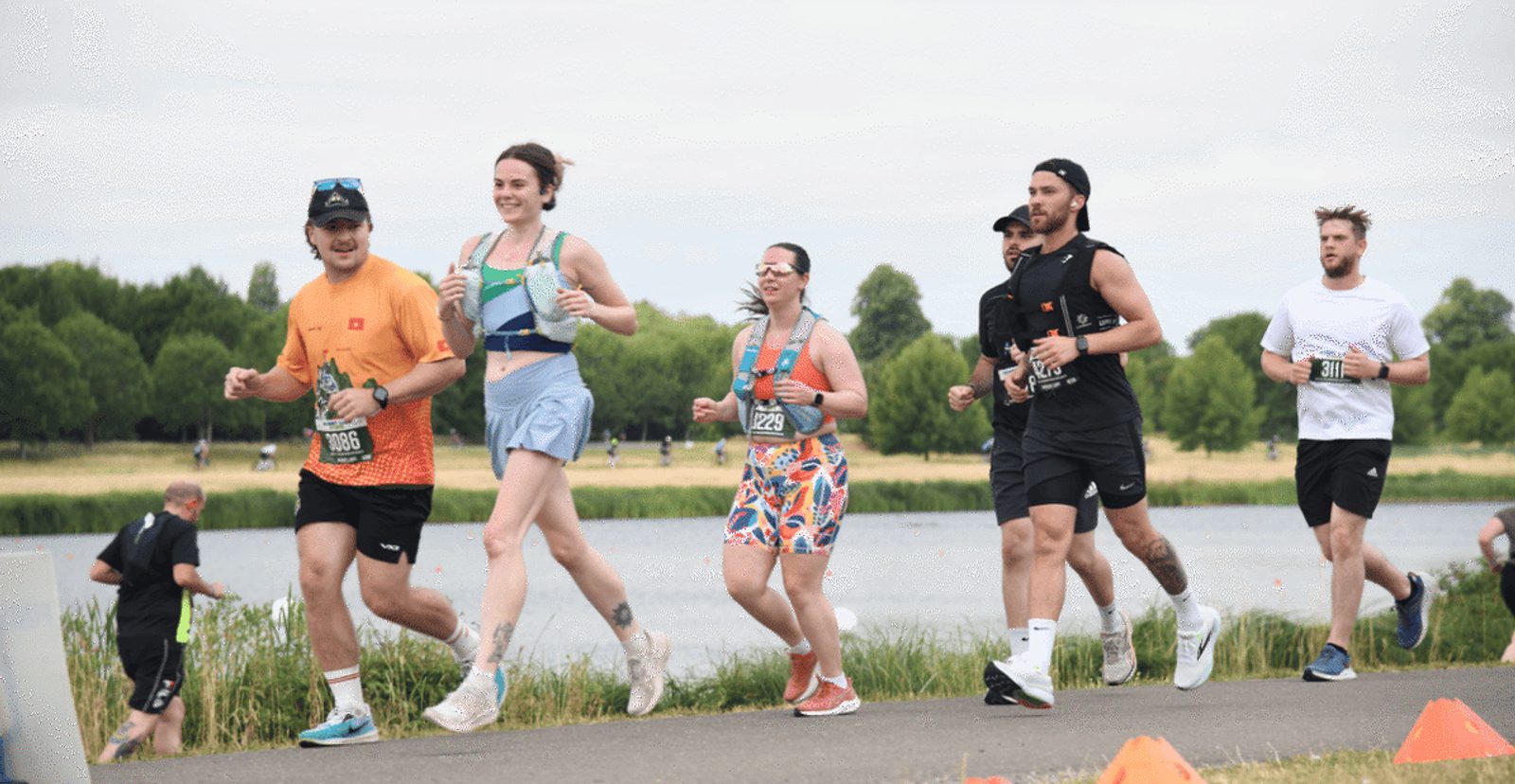 A group of runners, both men and women, jog along a paved path beside a lake in a park, wearing athletic gear and race bibs, with green trees and grass in the background.
