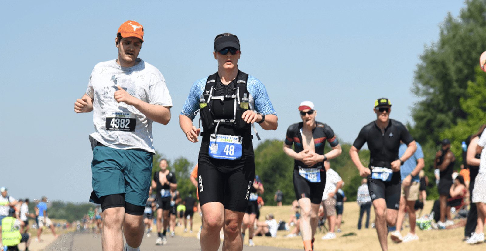A group of athletes runs outdoors during a race on a sunny day, with race bibs on their shirts and people watching from the sidelines.