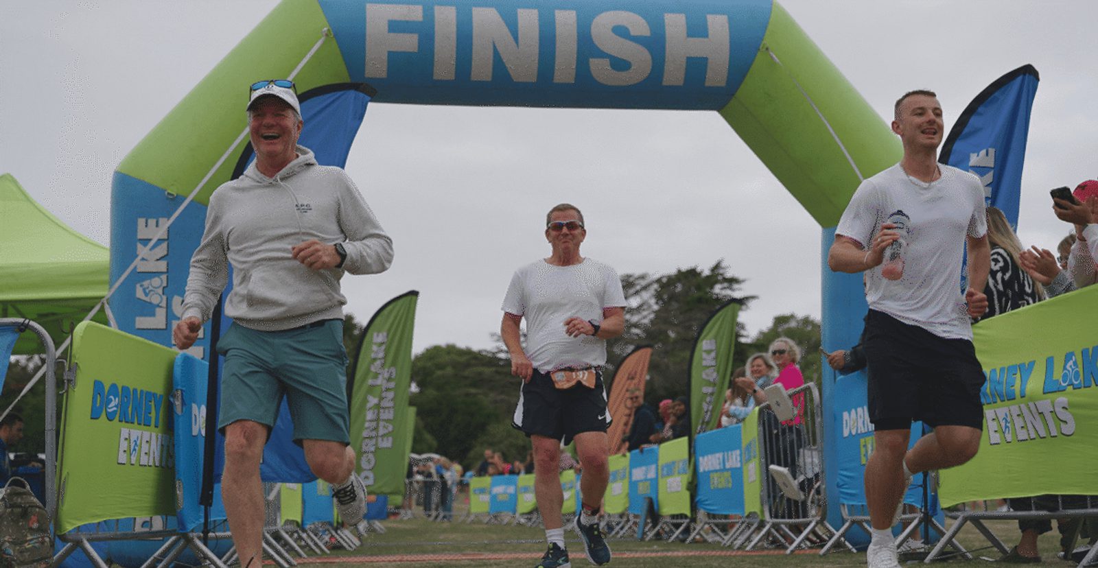 Three people in athletic wear smile and run as they cross a blue "FINISH" line at an outdoor event, with spectators and banners in the background.