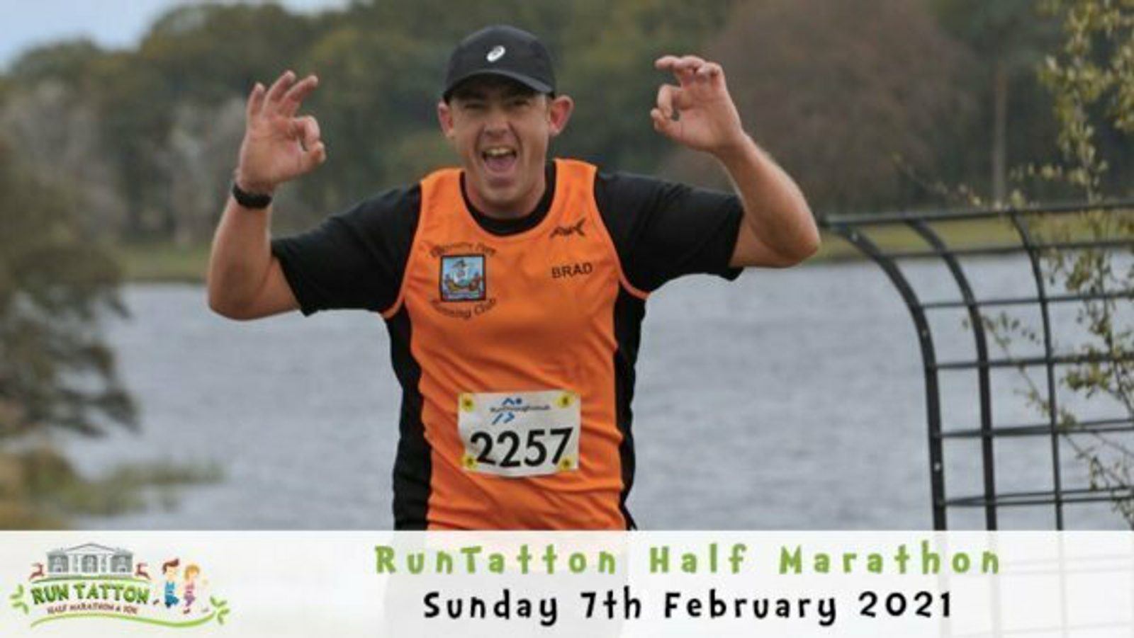 A runner in an orange and black shirt with the name "BRAD" and the number "2257" on his bib smiles and makes a gesture with both hands as he nears the finish line of the RunTatton Half Marathon. The event date is Sunday, 7th February 2021.