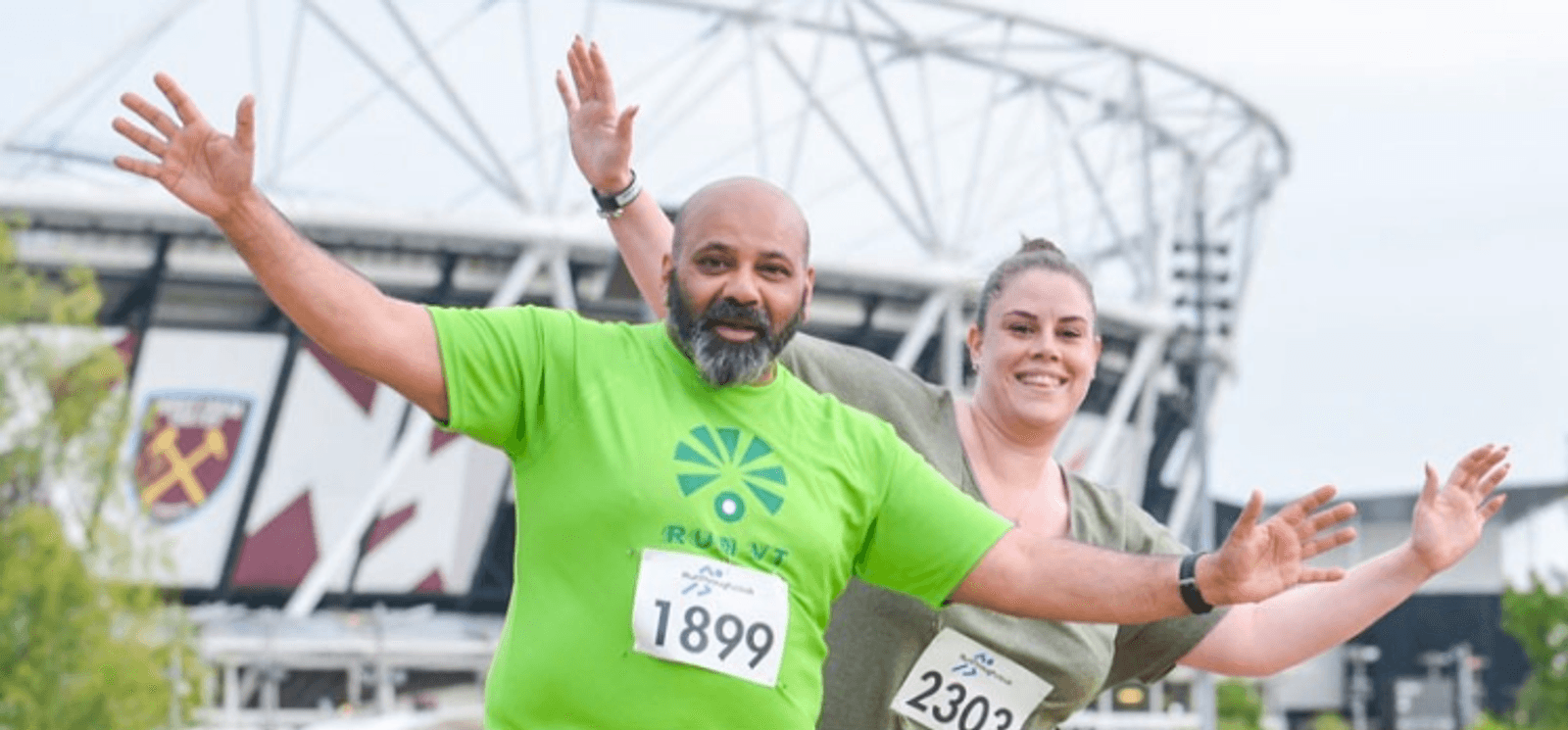 Two runners, a man in a green shirt with race number 1899 and a woman in a gray shirt with race number 2302, pose joyfully with outstretched arms in front of a stadium. Both are smiling, showing their enthusiasm and enjoyment of the event.