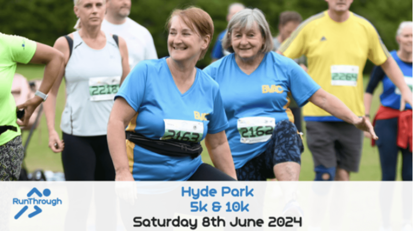 People participating in a group stretch at a running event in Hyde Park. Two women in front, wearing race bibs, are smiling and stretching their legs. The event banner reads "Hyde Park 5k & 10k, Saturday 8th June 2024.