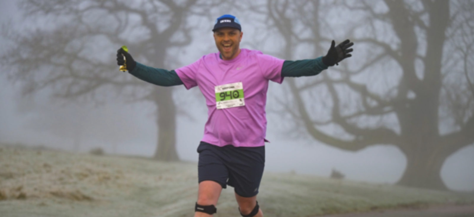 A runner dressed in a purple shirt, black knee braces, and dark shorts is jogging on a foggy path with open arms and a big smile. He is holding something in his right hand and wearing a blue cap. Tall, leafless trees and frosty grass are in the background.