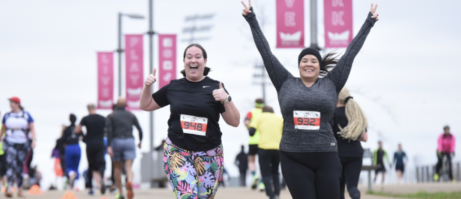 Two women, both wearing race bibs, smile and gesture as they run in an outdoor race. The woman on the left gives a thumbs up, and the woman on the right flashes a peace sign. Other runners can be seen in the background, and red banners hang from poles above the path.