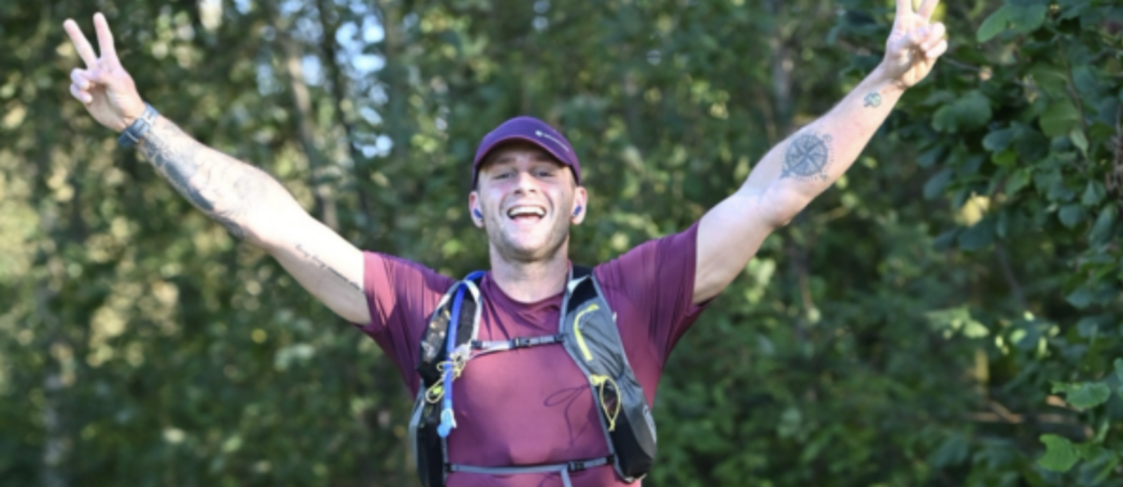A person dressed in a maroon shirt and cap, wearing a hydration pack, smiles broadly while raising both arms with peace signs. The person is standing outdoors against a backdrop of green trees.