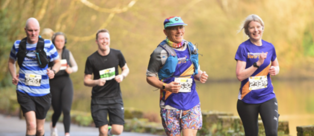 A group of runners participating in a race along a scenic outdoor path, surrounded by greenery. In the forefront, two women and one man are smiling and wearing race numbers. Other runners are visible in the background. The atmosphere is cheerful and energetic.