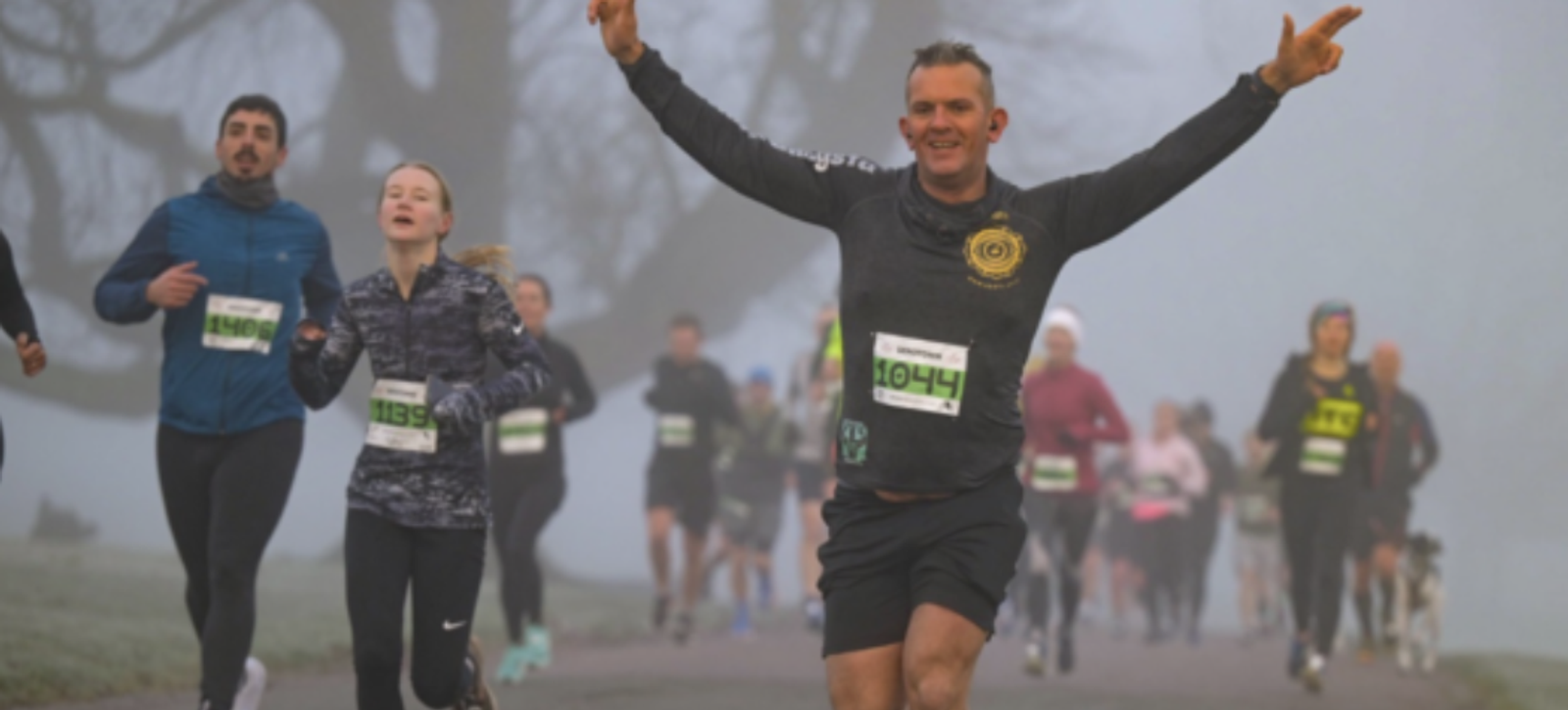 A group of runners participates in a race on a foggy day. In the foreground, a man in a dark shirt with a number "1044" is running with his arms raised in celebration. Other runners, wearing various athletic outfits and race numbers, follow behind him.
