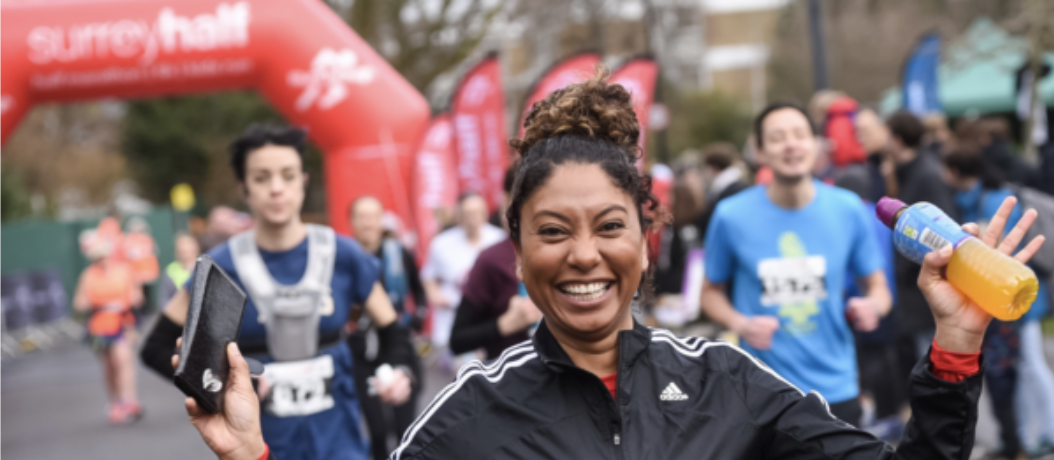 A smiling woman in athletic gear holds a smartphone and a bottle of orange drink while running in a race. Other runners are visible in the background under a red archway with the words "Surrey Half" on it. The atmosphere appears energetic and joyful.