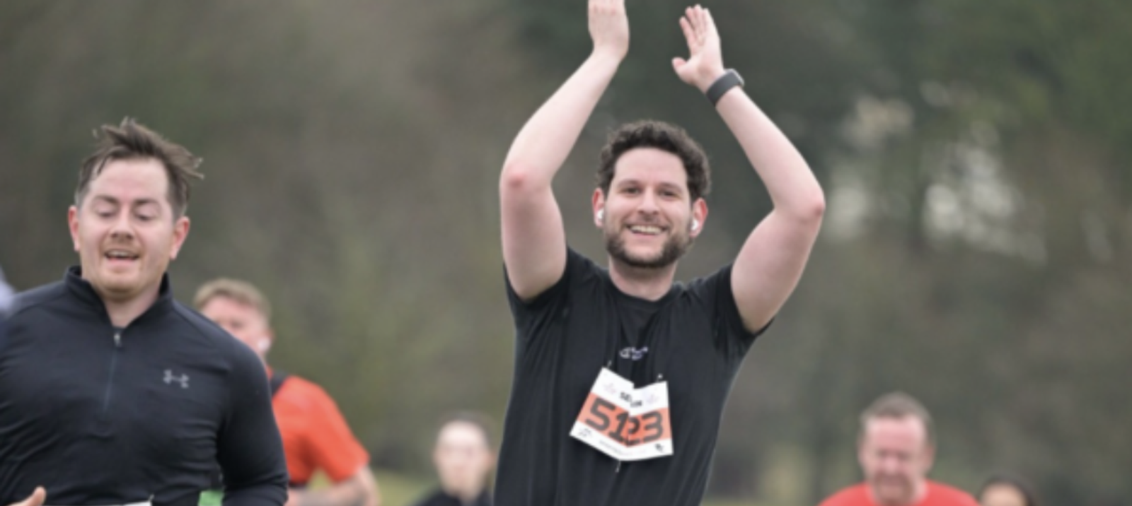 A group of runners participates in an outdoor race. A male runner in the foreground, wearing a black shirt and a number 523 bib, is smiling and clapping above his head. Other runners, blurred in the background, are focused on their strides with a mix of determination and effort.