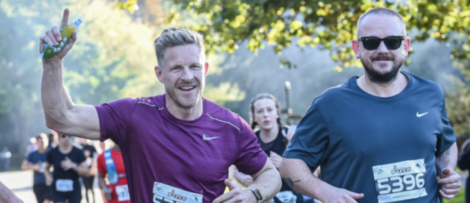 Two men are running in a race. The man on the left is wearing a maroon shirt and smiling while holding a drink bottle and raising one arm. The man on the right is wearing a dark shirt with sunglasses. Both have race bibs with numbers, and other runners are visible in the background.