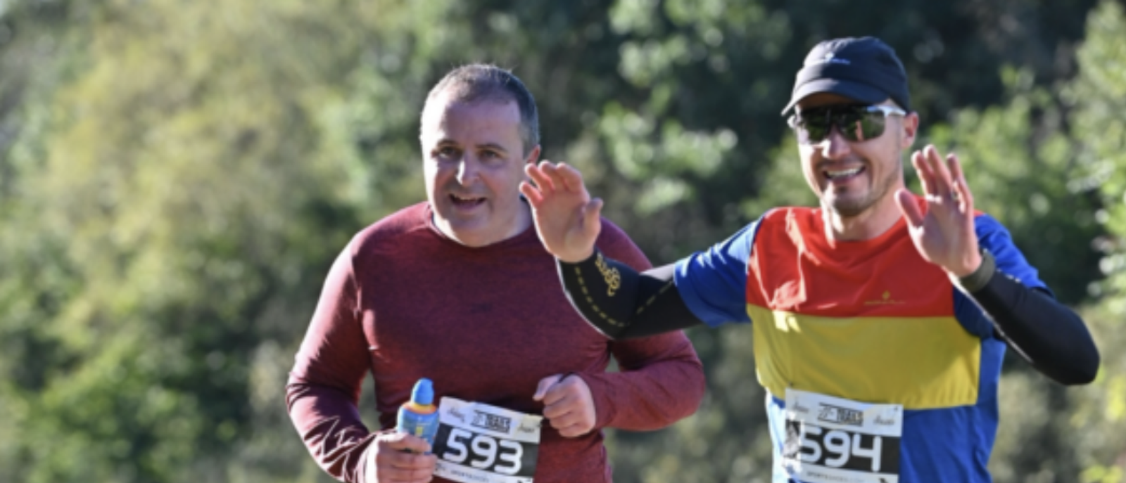 Two runners with race bibs numbered 593 and 594 are participating in a marathon or outdoor run. The runner on the left is wearing a red shirt and holding a water bottle, while the runner on the right, wearing a colorful shirt and sunglasses, waves and smiles. Trees and greenery are in the background.