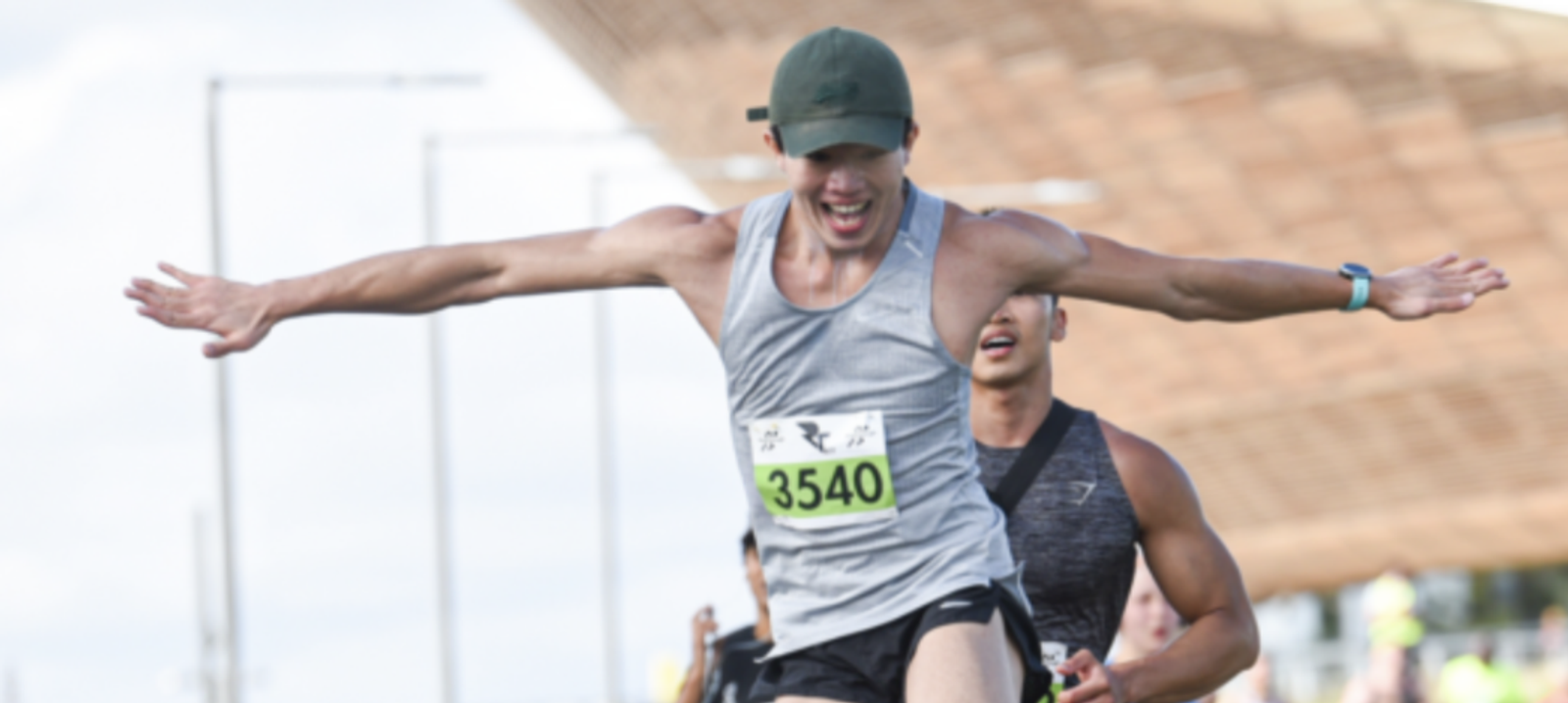 A runner wearing a gray tank top, black shorts, and a green cap with bib number 3540 is stretching his arms out in celebration as he finishes the race. Another runner is slightly behind him, both with joyful expressions. Urban background visible.