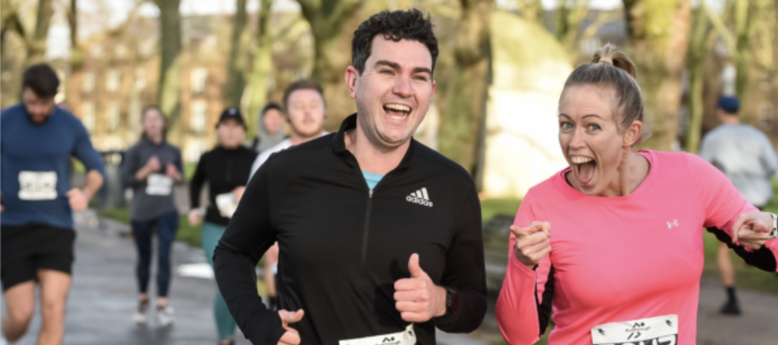 A group of runners participate in a race on a paved path lined with trees. In the foreground, two runners, one in a black long-sleeve shirt and another in a pink long-sleeve shirt, smile and gesture enthusiastically towards the camera.