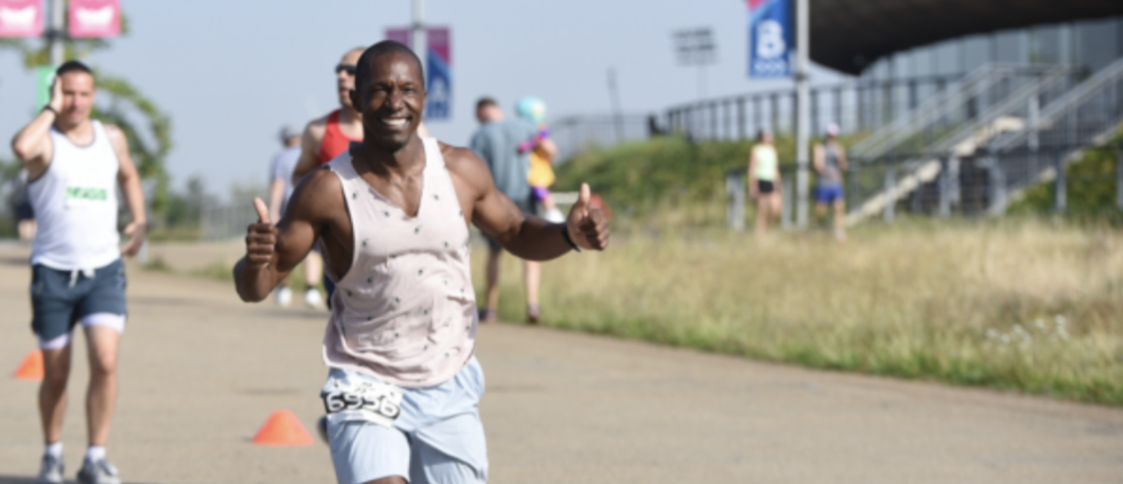 A smiling runner gives a double thumbs-up during a race. He is wearing a light pink sleeveless shirt and light blue shorts, and his race number is visible. Other runners and a grassy field with a stadium in the background can also be seen.