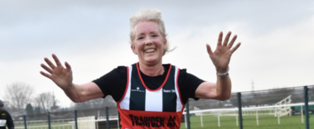 An older woman with short gray hair is smiling and raising her hands as she runs. She is wearing a red and black race vest with "TEAINCHECK" written on it. The background shows a sports field with a cloudy sky.