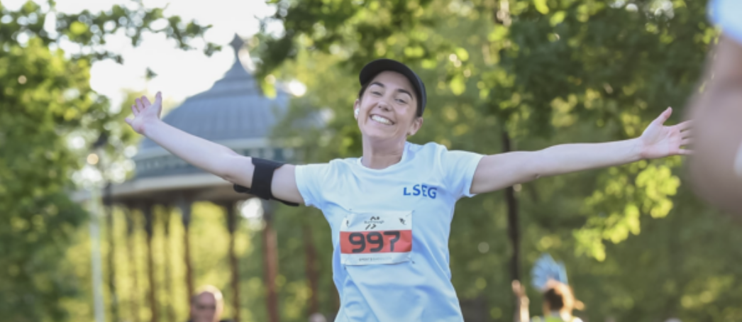 A person wearing a light blue shirt and a black cap smiles broadly with arms outstretched while running in a park. The runner's race number 997 is visible on their shirt. Green trees and a gazebo are in the background, suggesting a sunny day.
