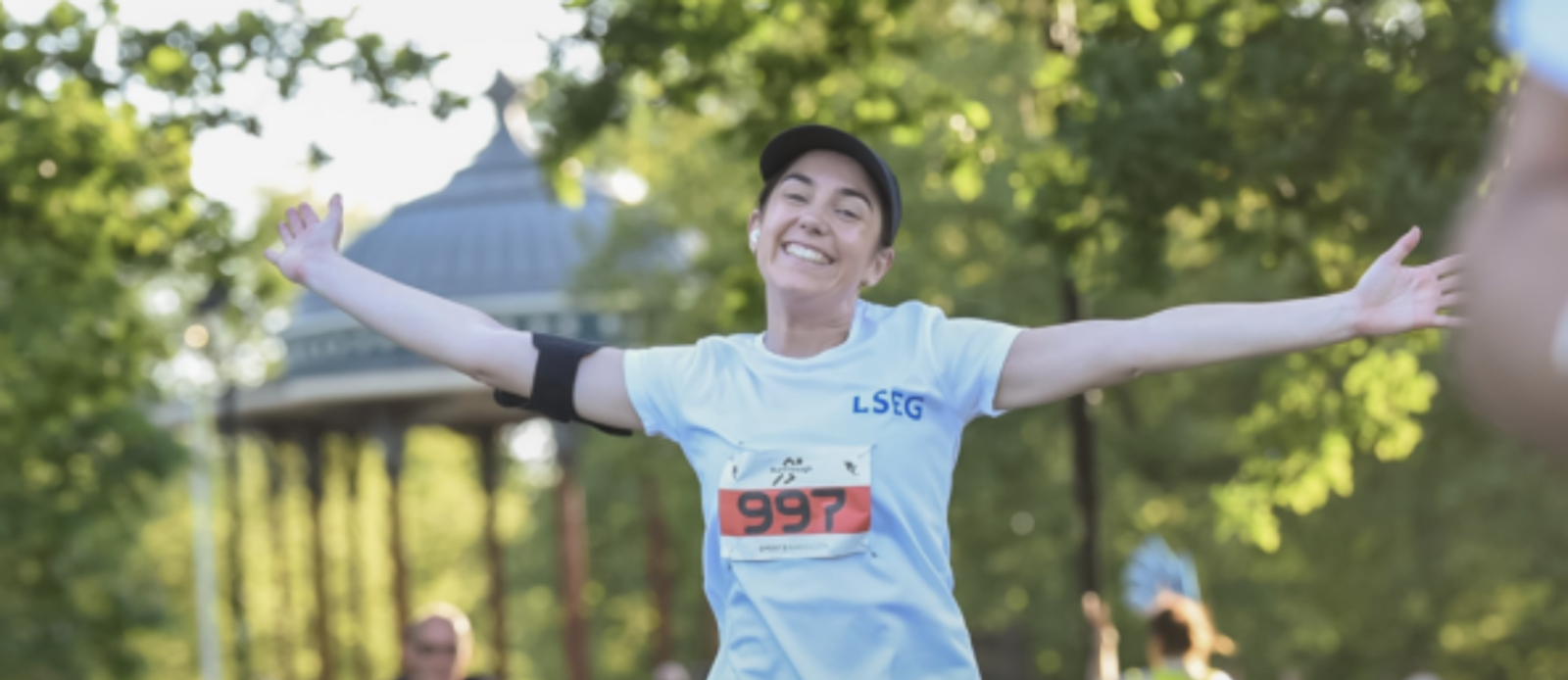 A person wearing a light blue shirt and a black cap smiles broadly with arms outstretched while running in a park. The runner's race number 997 is visible on their shirt. Green trees and a gazebo are in the background, suggesting a sunny day.