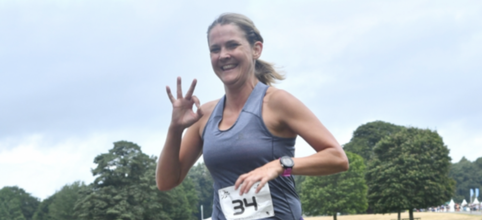 A smiling runner wears a grey tank top and a race bib numbered 34. She gestures with her right hand while running outdoors on a cloudy day. Green trees are visible in the background.