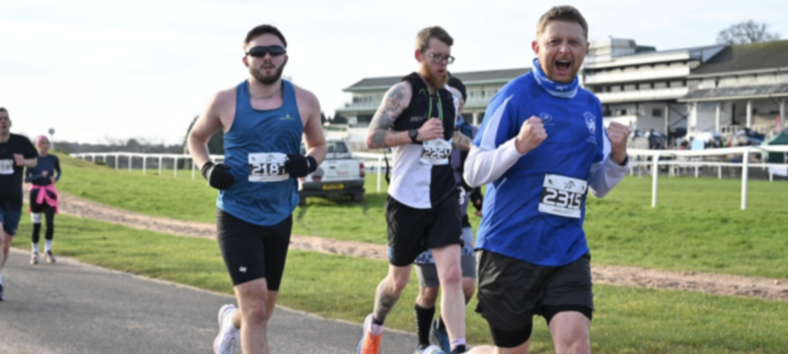A group of runners participates in a race on a paved path adjacent to a green field. The front runner in a blue shirt and number 2315 is cheering with his fist in the air. Other runners behind him appear focused. A large building is visible in the background.