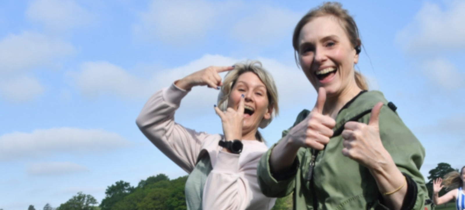 Two women smiling and posing playfully outdoors with a blue sky and scattered clouds in the background. The woman on the left is making a peace sign with her hand, and the woman on the right is giving a double thumbs-up gesture. Green trees are visible behind them.