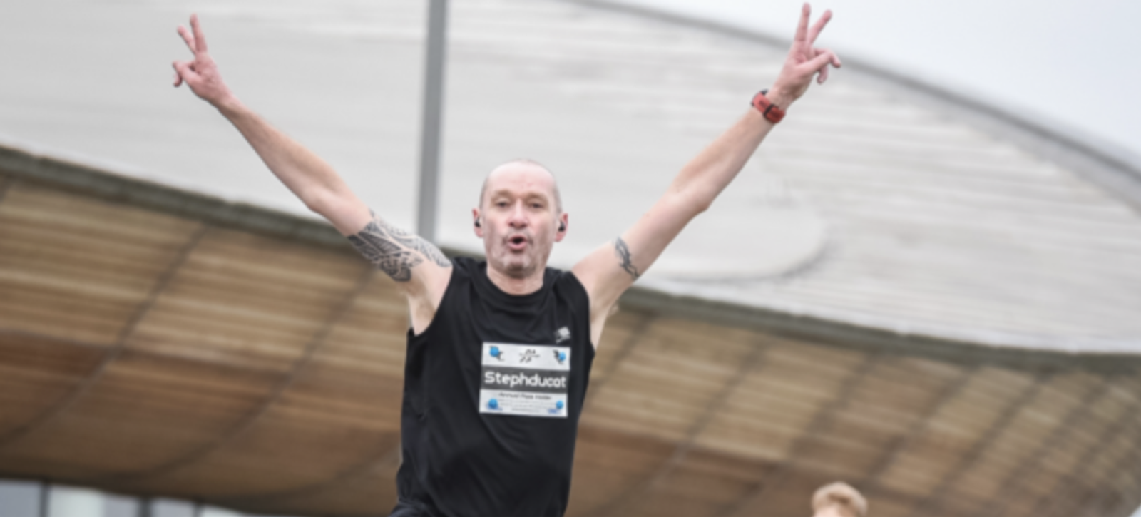 A runner with tattoos and a black tank top, bearing a race bib with "Steph" visible, raises both arms in a victory gesture, making peace signs with both hands. He is outdoors with a large, curved building structure in the background.