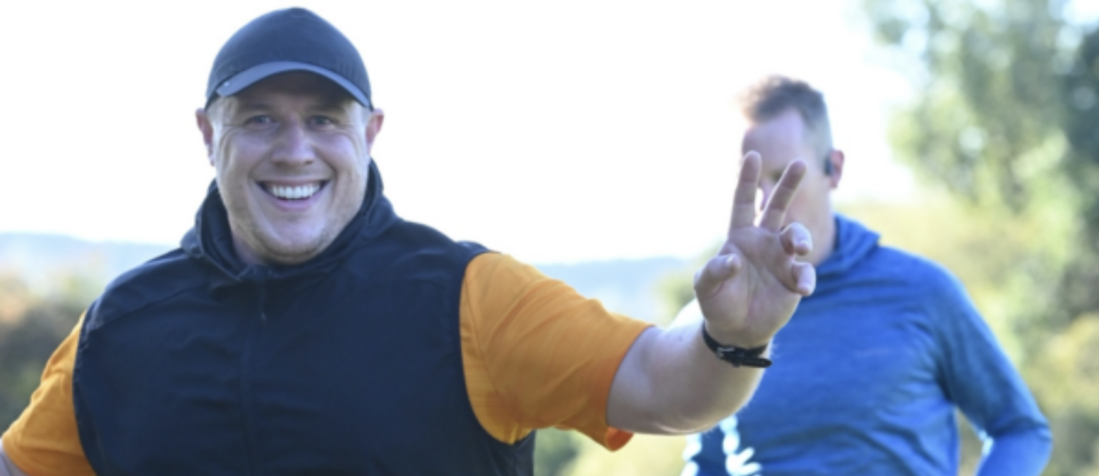 A smiling man wearing a black cap, black vest, and orange shirt holds up a peace sign while jogging outdoors. Another person in a blue shirt jogs in the background. It is a sunny day with trees and greenery in the distance.