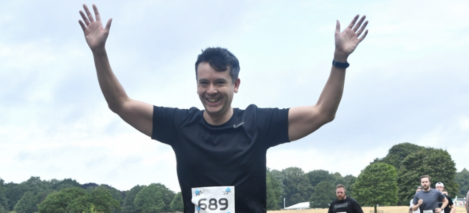 A man wearing a black shirt with race bib number 689 is smiling and raising his arms in celebration. He is outdoors, with a few other runners in the background and a cloudy sky overhead. Green trees can be seen in the distance.