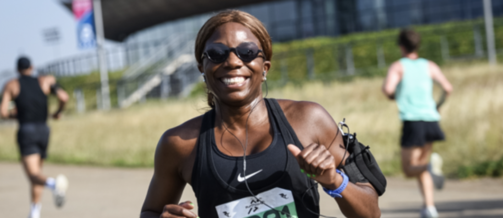 A woman with dark skin and long hair is running in a race outside. She is wearing black athletic wear, sunglasses, and earphones. She has her race number pinned to her shirt and is smiling. Other runners are also visible in the background.