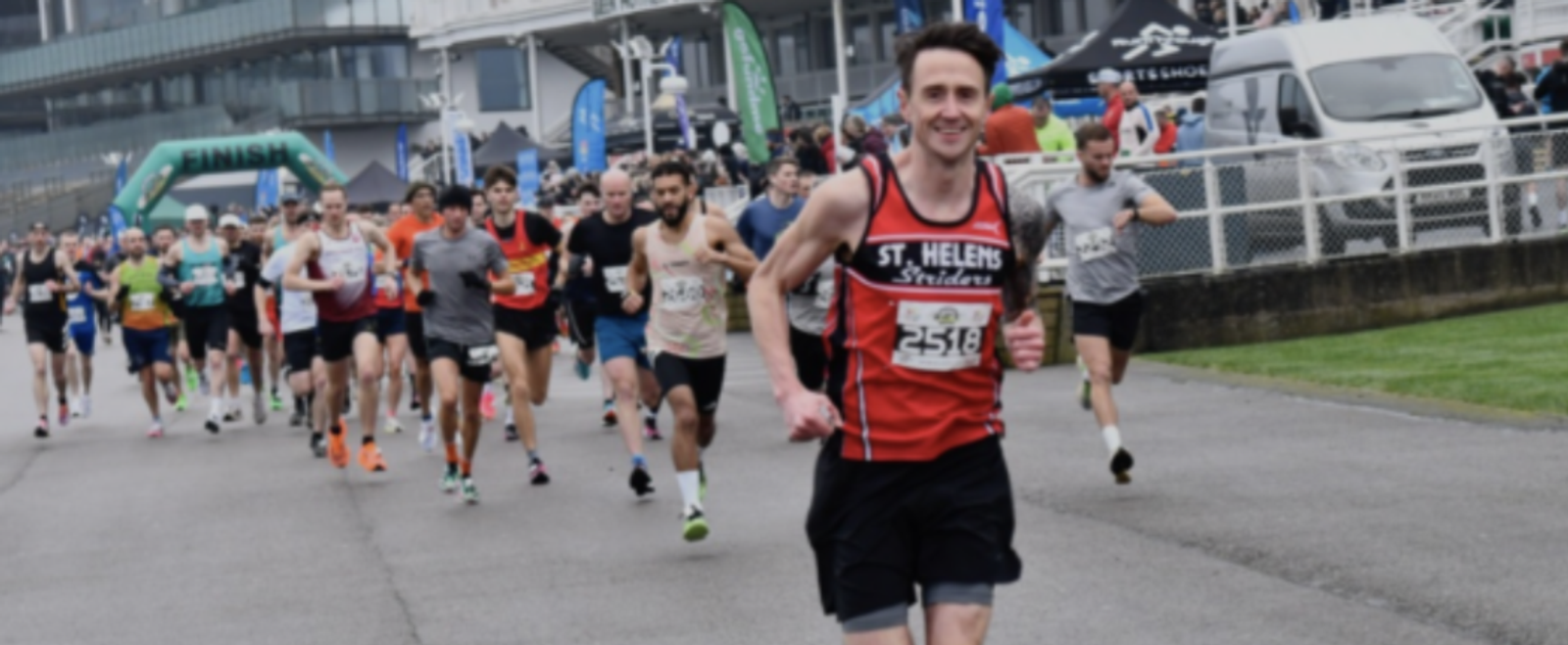 A group of runners competing in a race on a cloudy day. The foreground shows a smiling runner wearing a black and red "St. Helens Striders" jersey and bib number 2516. The finish line is visible in the background along with other participants and spectators.