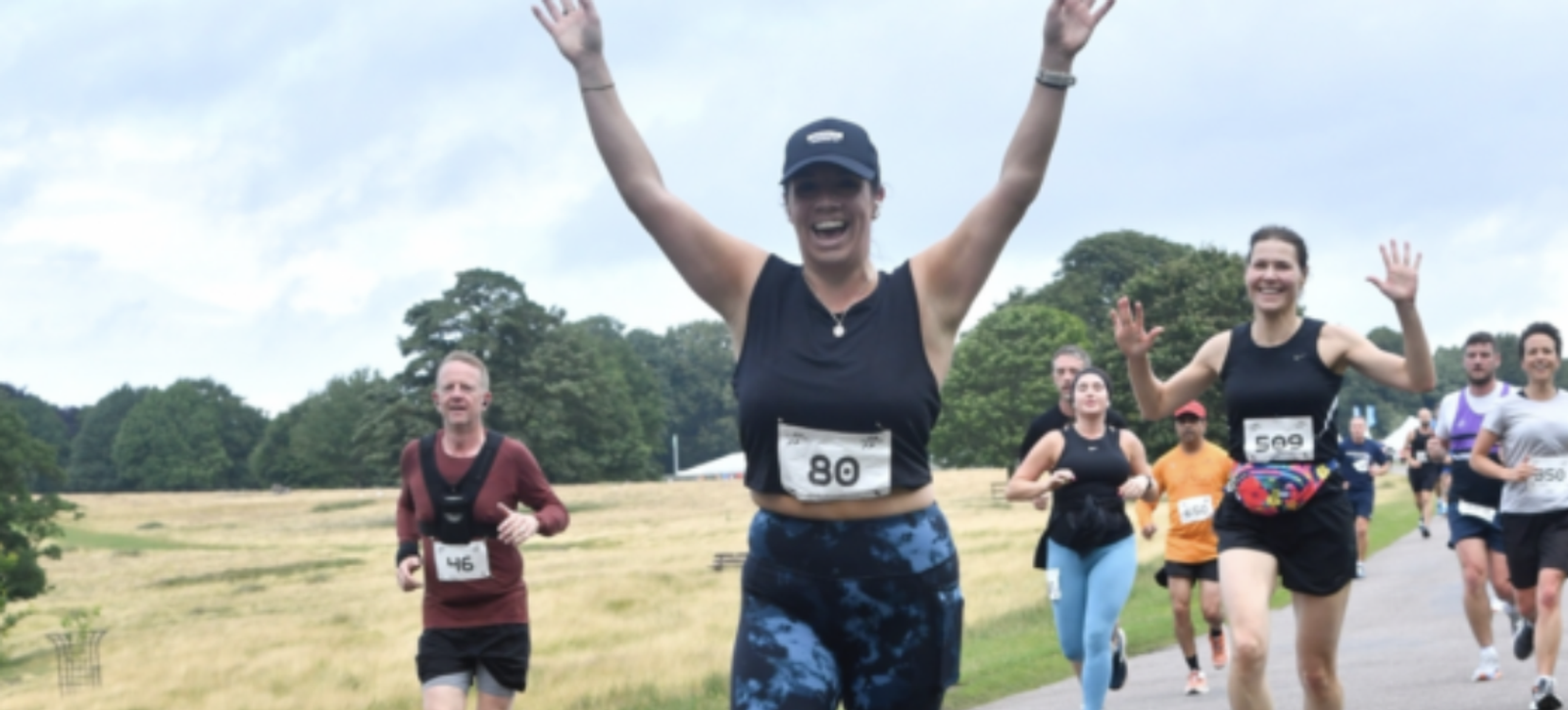 A group of runners is participating in a race on a paved path in a park. The runner in the foreground, wearing a black cap and tank top with blue shorts, raises her hands in celebration, while others follow behind, some also raising their hands and smiling. Trees and grass surround the participants.