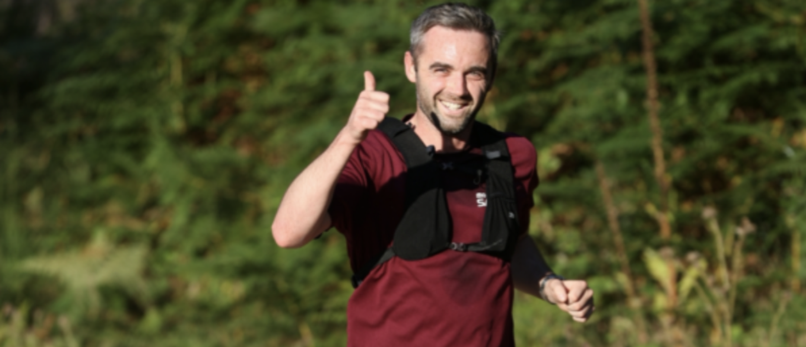 A man wearing a maroon shirt and a black running vest gives a thumbs-up while jogging. He is smiling and appears cheerful, with trees and greenery blurred in the background.