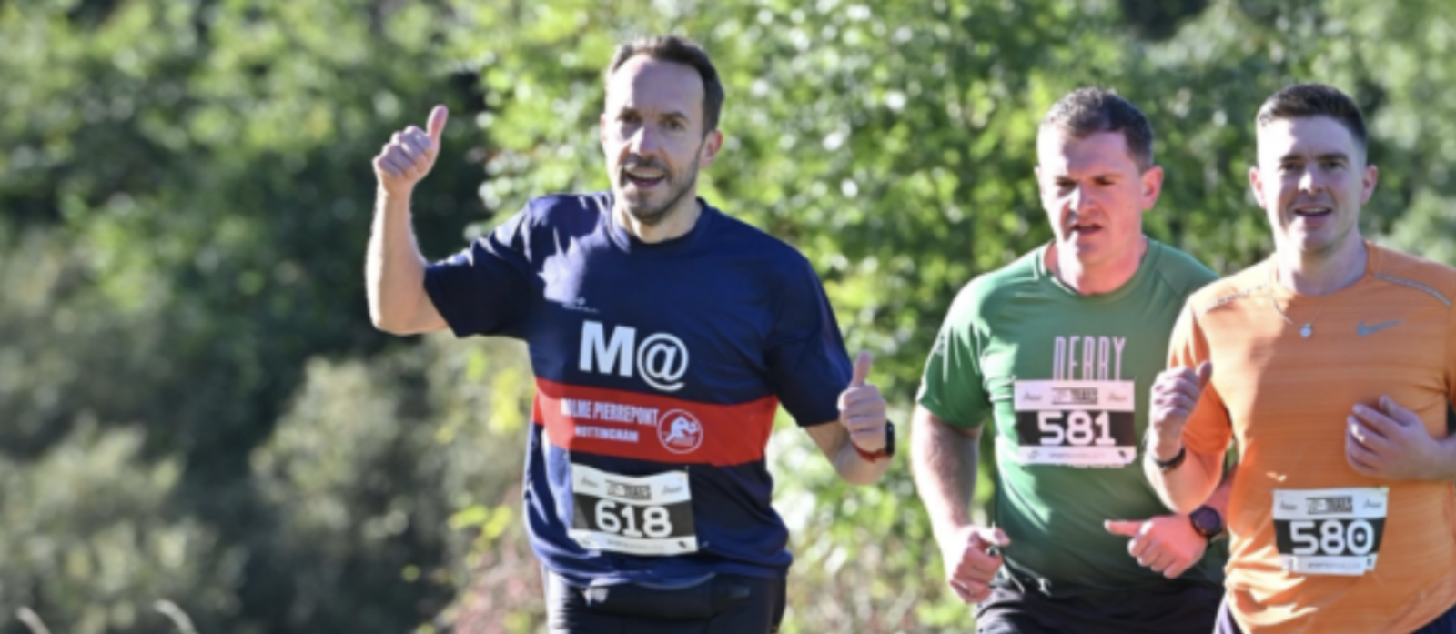 Three men in athletic attire are running outdoors. One man wears a blue shirt and gives a thumbs-up. Another wears a green shirt with "Derby" on it, and the third wears an orange shirt. They all have bib numbers: 618, 581, and 580, respectively. Trees are in the background.
