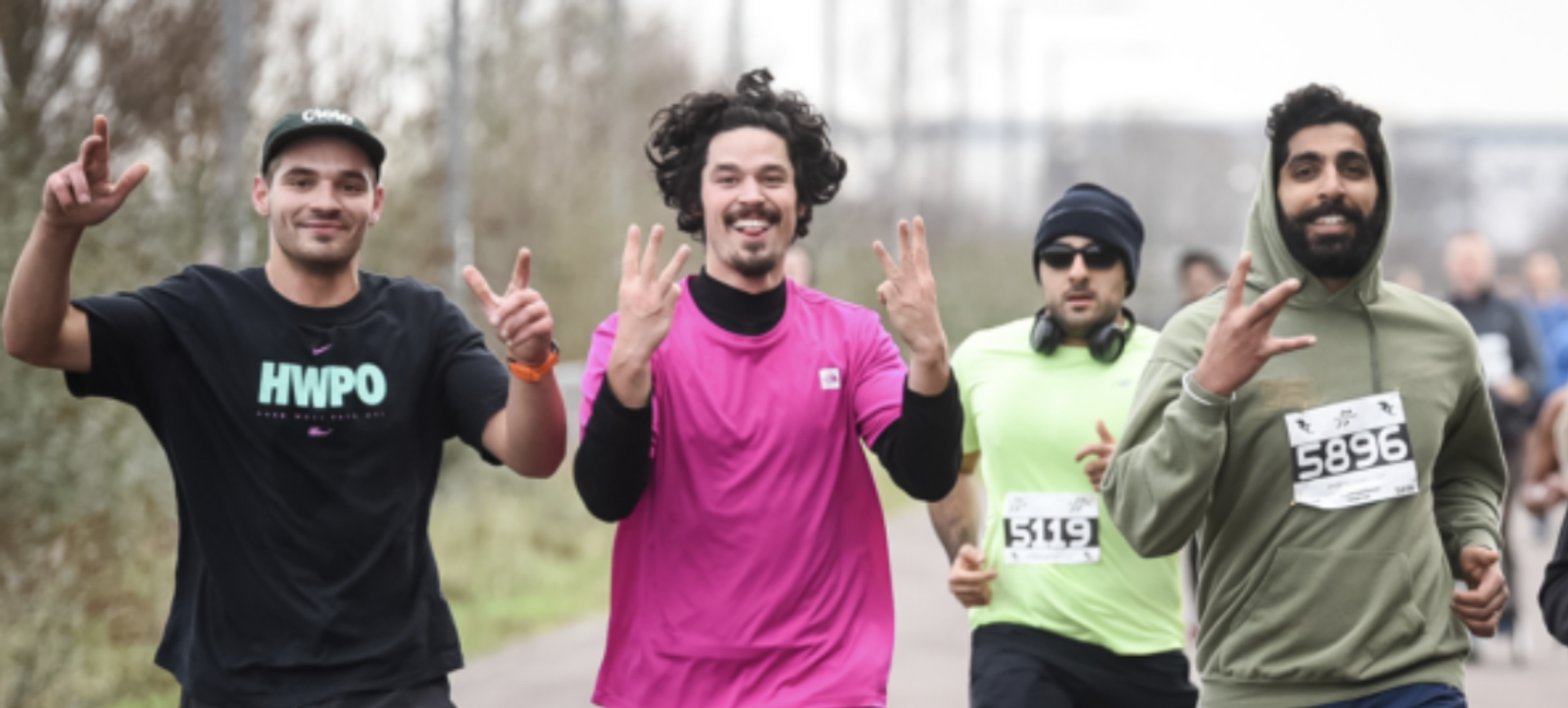 Three men participate in a race, smiling and making hand gestures at the camera. The man on the left wears a black shirt, the man in the middle wears a pink shirt, and the man on the right wears a green shirt and a race bib number 5896. Another runner is partly visible behind them.