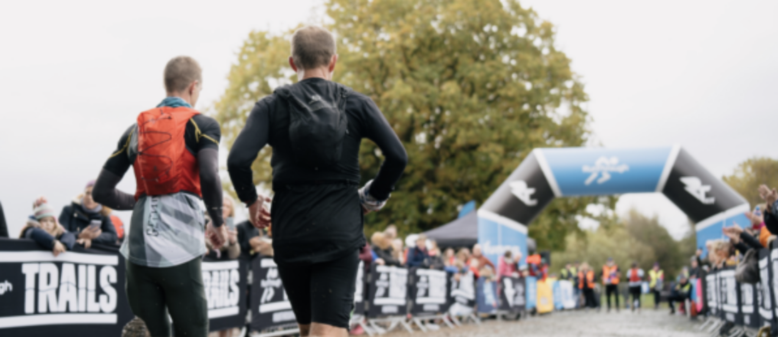 Two runners in athletic gear race towards the finish line at an outdoor event. The path is muddy and lined with cheering spectators behind barrier banners that read "TRAILS." A large inflatable arch, marking the finish line, stands in the background under a cloudy sky.