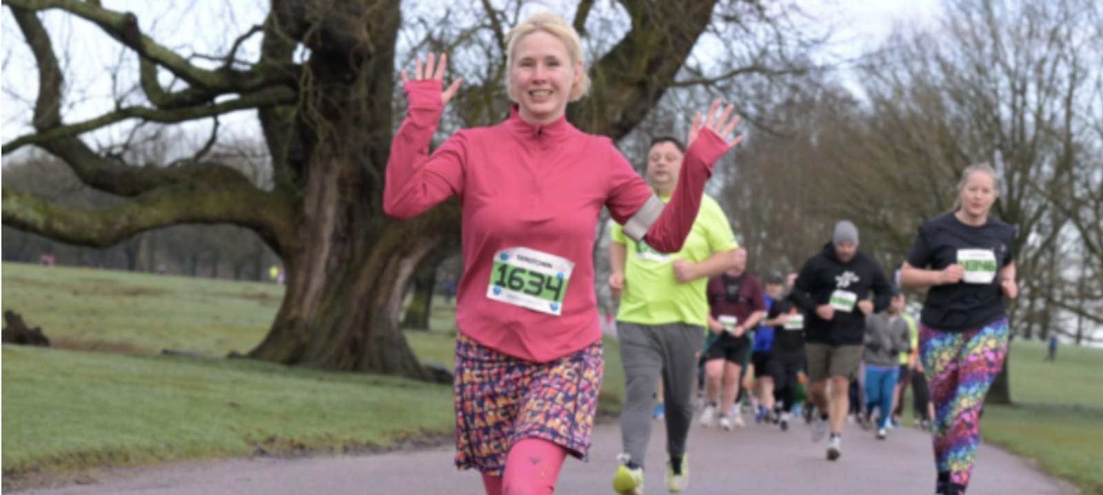 A woman wearing a bright pink running outfit with a race number 1654 pinned to her shirt is smiling and waving as she runs along a park path. Behind her, several other runners dressed in various colorful outfits follow. Trees and a grassy area surround the path.
