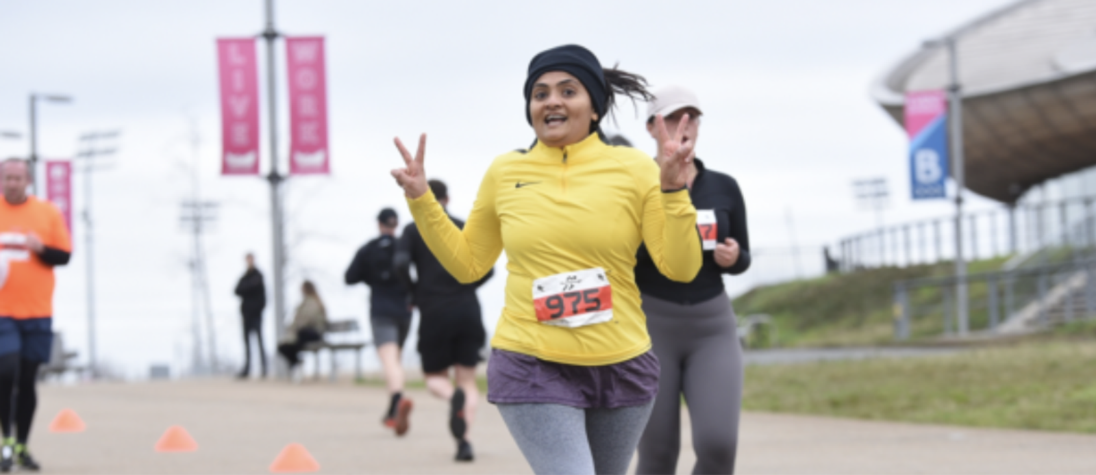 A woman in a yellow jacket and purple shorts is running in a race, smiling and making a peace sign with both hands. She is wearing a race number 975. Other runners can be seen in the background, with pink banners on poles lining the path.