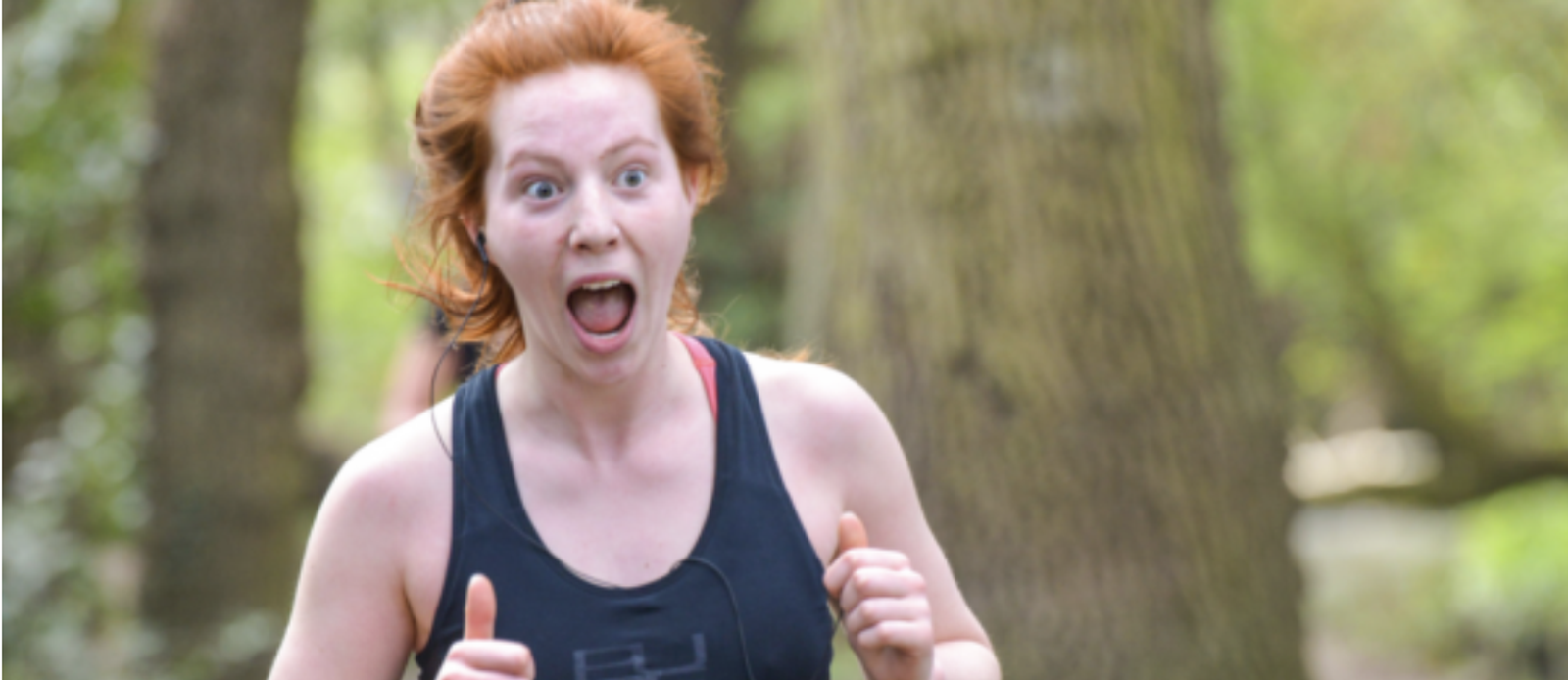 A person with red hair wearing a black tank top is running in a forested area with a surprised or excited expression on their face. Their mouth is open and their eyes are wide, with trees and greenery in the background.