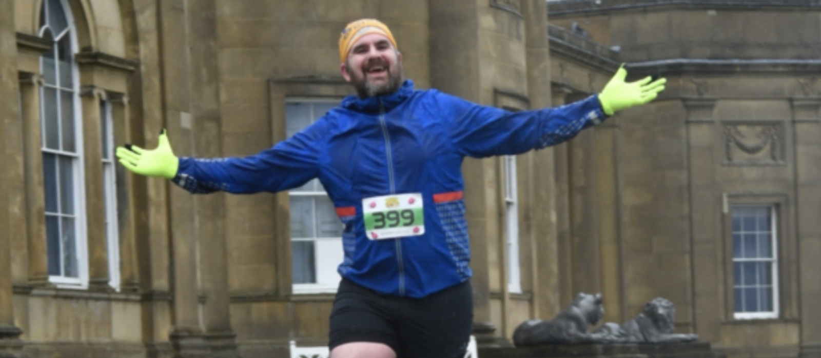 A person wearing a blue jacket, black shorts, a yellow headband, and fluorescent yellow gloves jubilantly extends their arms while running. The bib number 399 is pinned to their chest. They are in front of a large stone building with ornate architectural details.