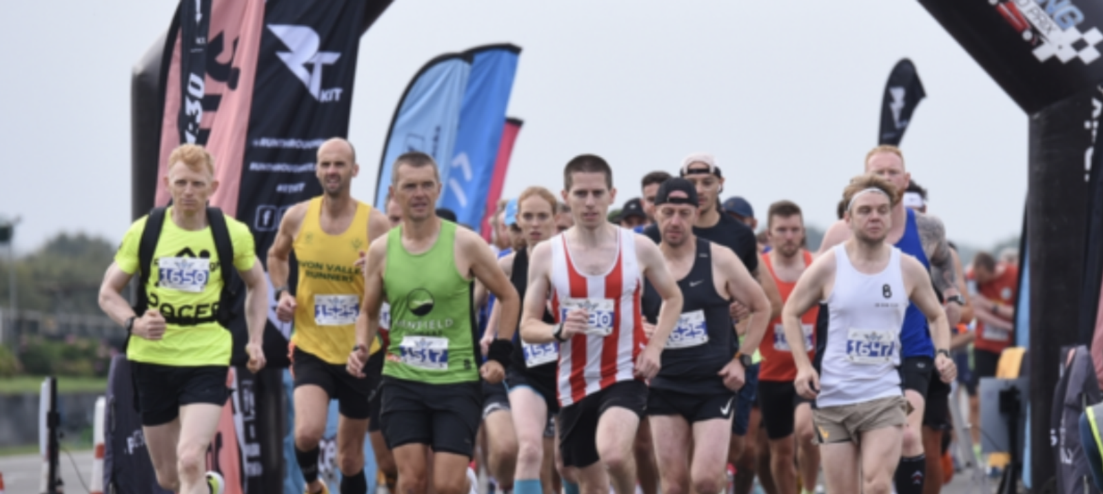 A diverse group of runners begins a race under an archway. Participants wear various athletic outfits with race numbers pinned to their shirts. Some wear headbands or hats. Colorful event flags and spectators can be seen in the background.