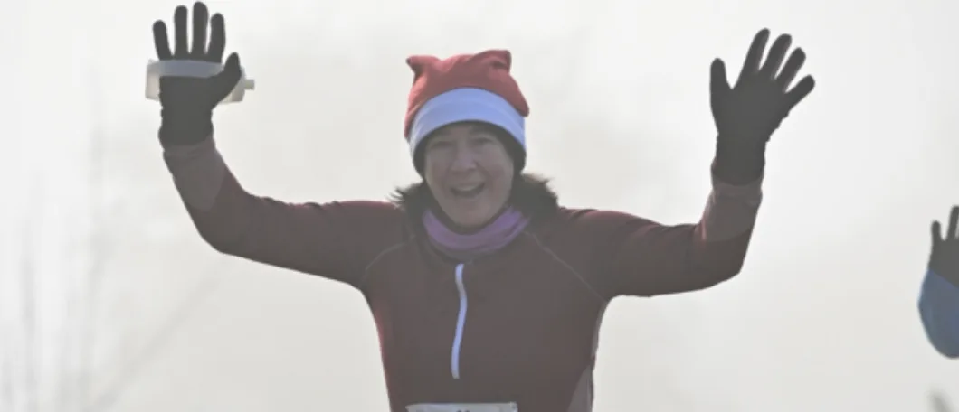 A person wearing a Santa hat, maroon jacket, and black gloves is joyfully raising both arms while smiling during a race or outdoor event. The background is foggy, adding a festive, wintry atmosphere to the scene.