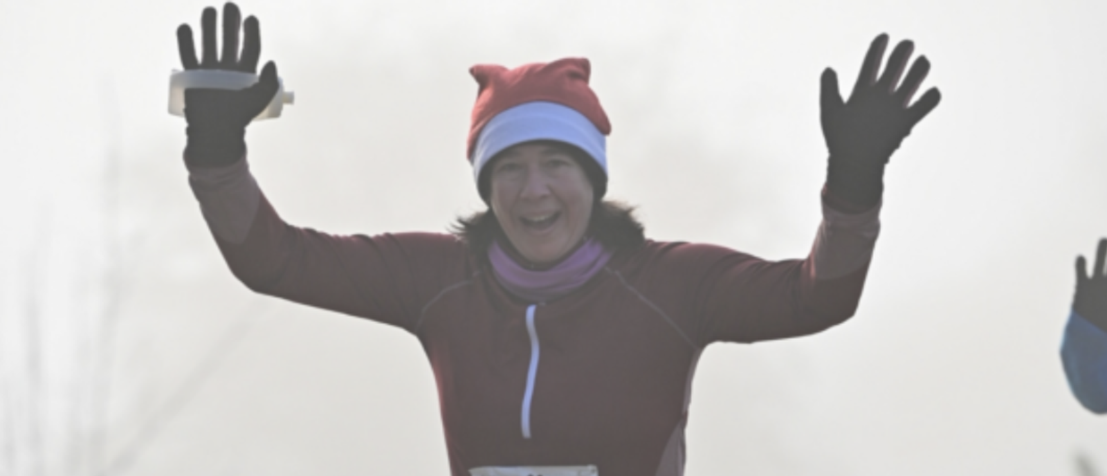 A person wearing a Santa hat, maroon jacket, and black gloves is joyfully raising both arms while smiling during a race or outdoor event. The background is foggy, adding a festive, wintry atmosphere to the scene.
