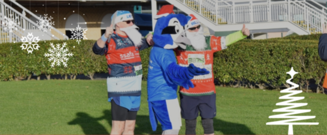 Three people, one in a blue mascot costume, wear festive outfits and Santa beards. Two are in red and green attire with printed logos. They pose with thumbs up and playful gestures against a backdrop of holiday decorations, including snowflakes and a white Christmas tree.