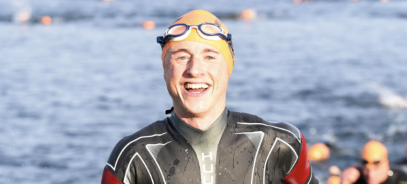 A triathlete in a wetsuit, orange swim cap, and goggles smiles broadly as he emerges from the water during a race. In the background, other swimmers can be seen in the water, with orange buoys marking the course.