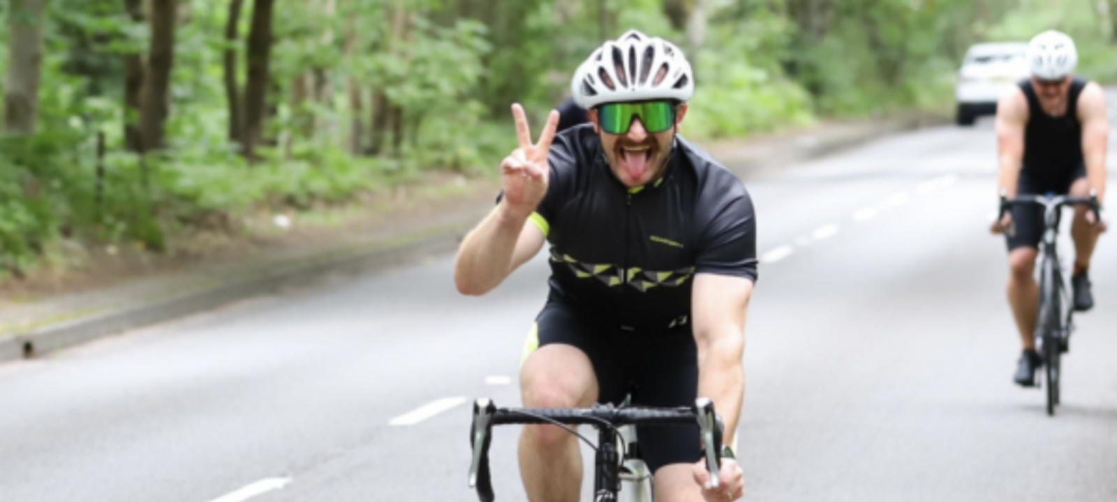 A cyclist in a black outfit and white helmet rides on a road, making a peace sign and sticking his tongue out. He is wearing large green sunglasses. Another cyclist can be seen in the background riding on the same road. Trees line the sides of the road.