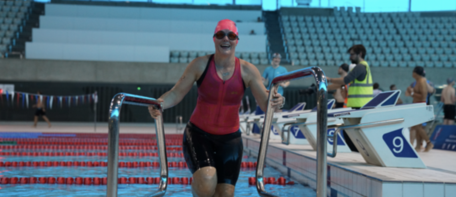 A smiling swimmer wearing a red swim cap and suit climbs out of an indoor pool using a metal ladder. The pool has multiple lanes marked by red lane dividers. In the background, spectators and officials are visible in the stands and pool deck.
