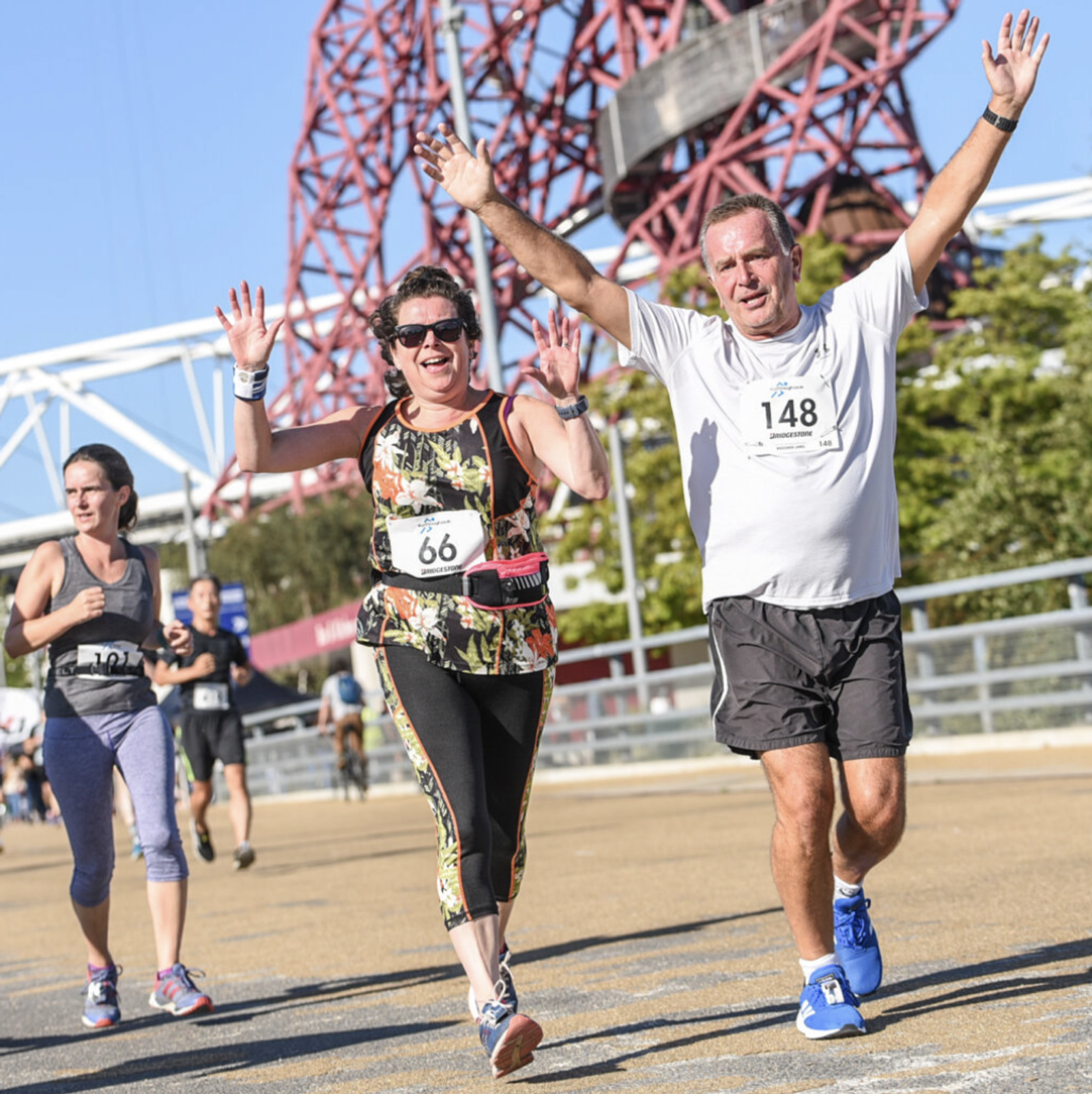 People running a race on a sunny day, raising their arms in celebration as they approach the finish line. A woman wearing colorful athletic clothes and a man in white with a race number are in the foreground. Tall, red iron structure and trees in the background.