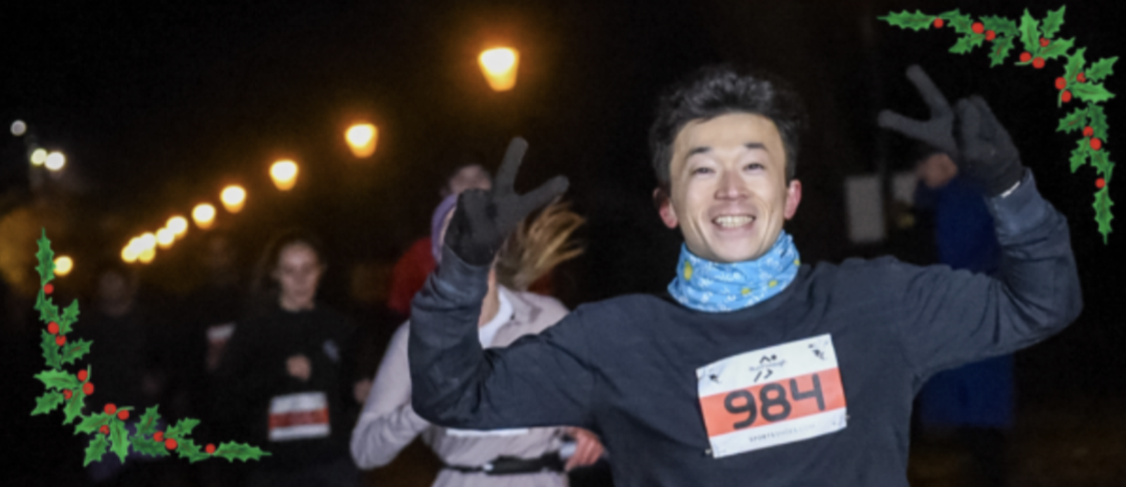 A man wearing a race bib numbered 984 is running at night. He is smiling widely and holding up both hands in a peace sign. Other runners are visible in the background. The image is decorated with holly branches in the top left and bottom right corners.