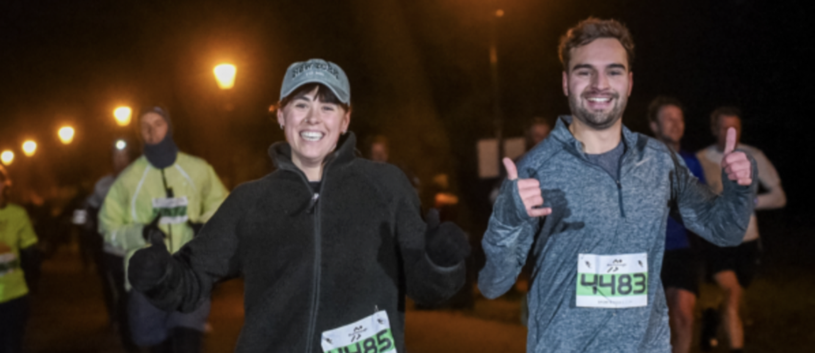 Two runners smile and give thumbs up as they run a nighttime race. Both wear numbered race bibs and warm, dark clothing. Streetlights illuminate the path, and several other runners are visible in the background. The atmosphere is lively and energetic.
